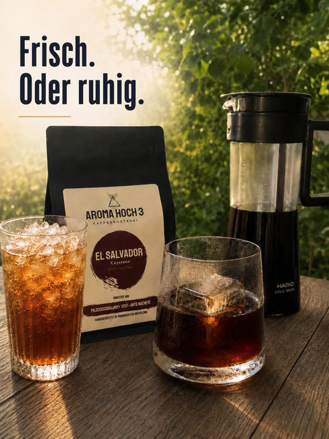 Iced Coffee mit Crushed Ice und Cold Brew mit großem Eiswürfel neben Aroma Hoch 3 El Salvador Kaffeepackung im warmen Sommerlicht